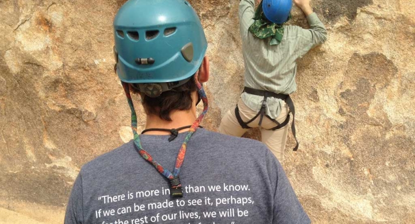 From behind, a person wearing a helmet watches another person who is also wearing safety gear scale a rock wall. 